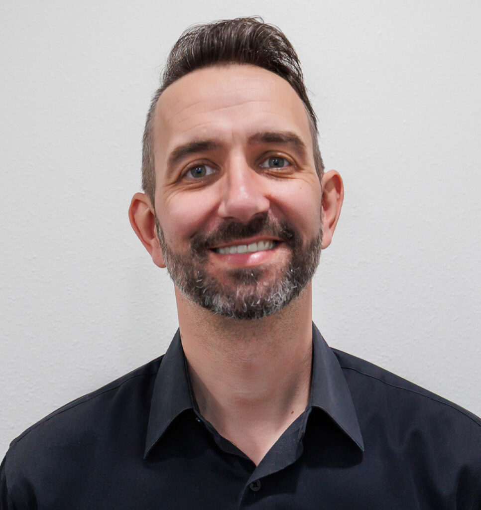Smiling man with short dark hair and gray beard wearing a black collared shirt against a light background.