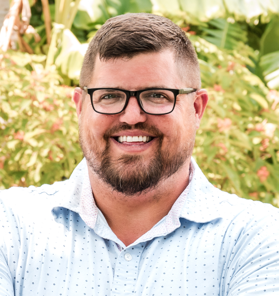 Smiling man with short hair and beard, glasses, light blue patterned shirt outdoors.