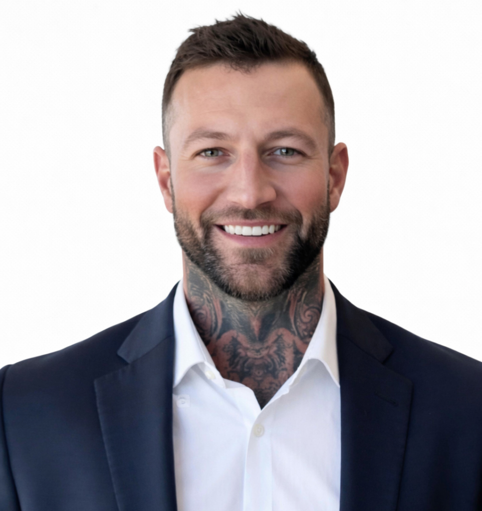 Smiling man with tattoos on neck, white shirt, dark blazer, white background.