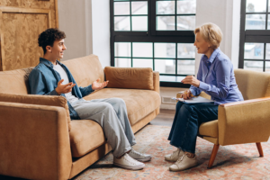 Young man talks with a woman in a beige living room, seated on a sofa and chair.