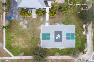 Aerial view of a fenced yard with a central basketball court, surrounding grass and trees.