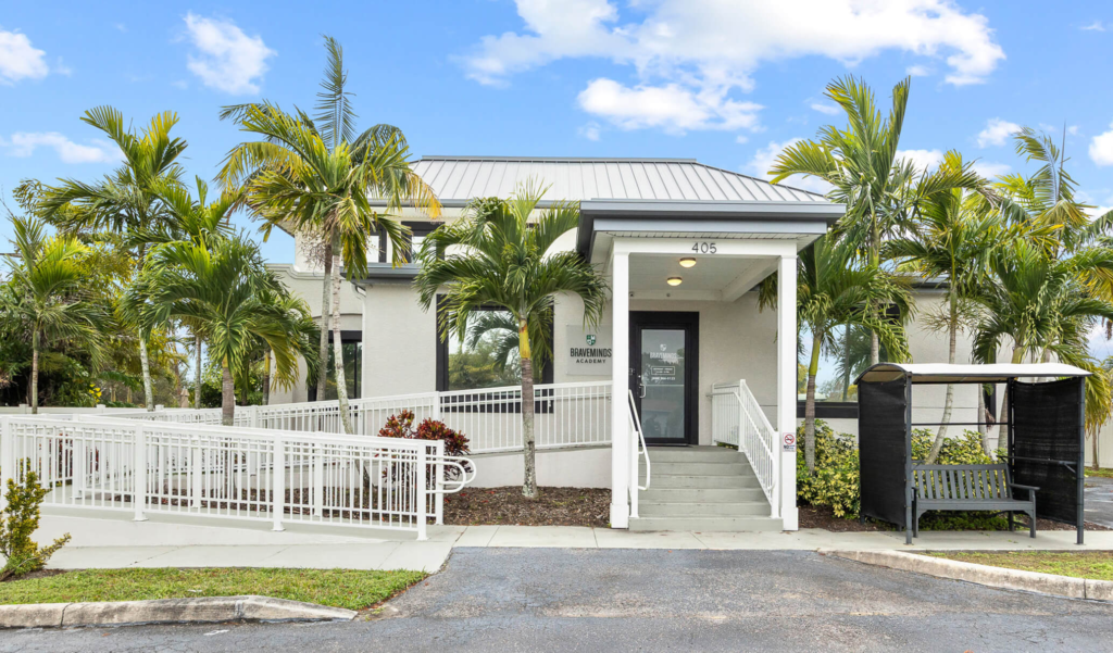 Braveminds Academy building with metal roof, glass doors, white fence, and palm trees.