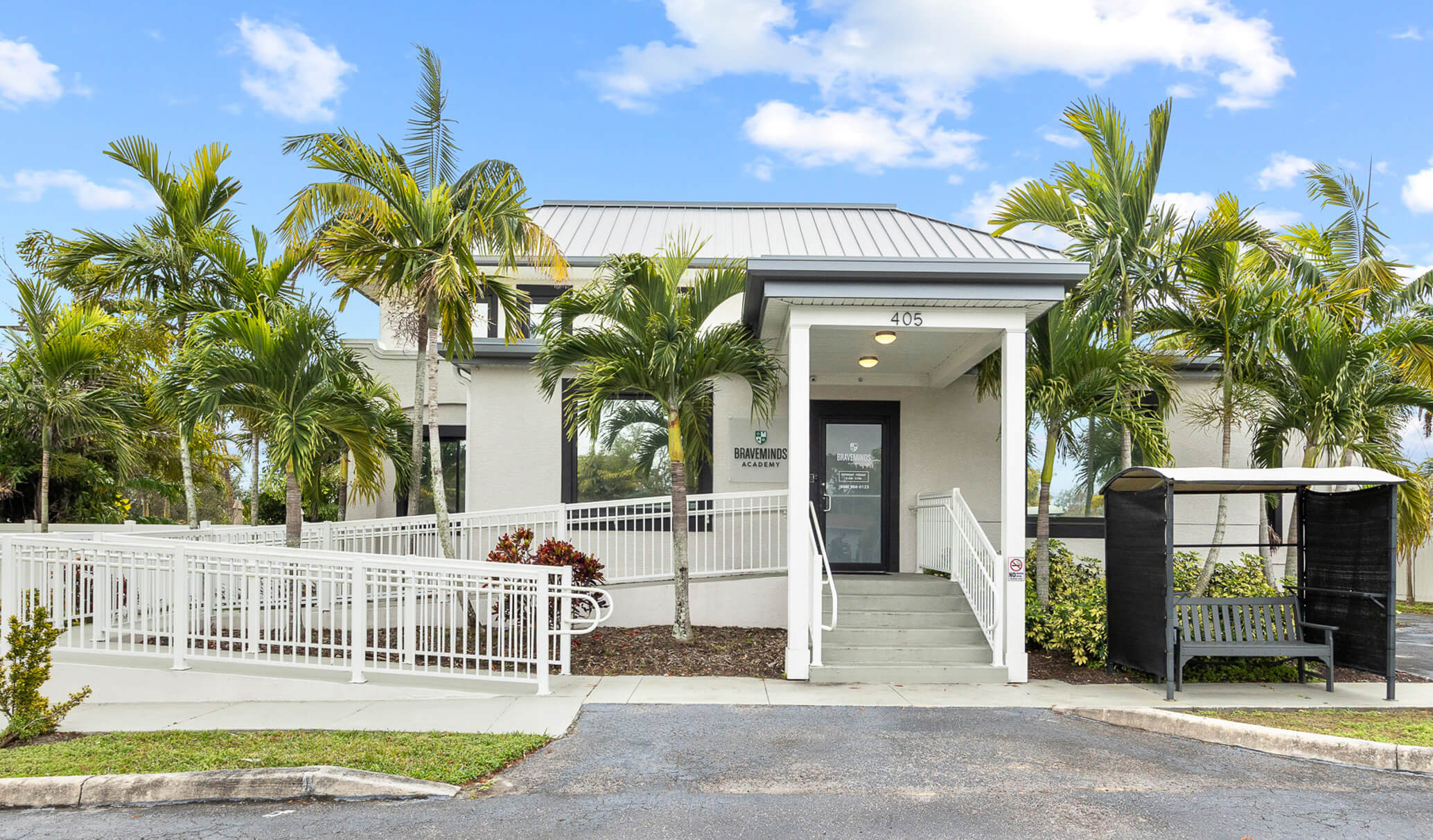 Braveminds Academy building with metal roof, glass doors, white fence, and palm trees.