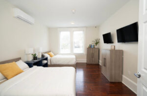 Bright bedroom with two single beds, white bedding, yellow pillows, and blue nightstands. TVs on wall. Wood floor.