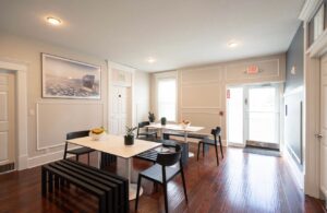 Bright communal dining area with white walls, dark wood floors, black furniture, and long tables.