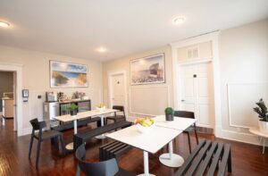 Bright communal dining area with white walls, wooden floors, tables, and black chairs.