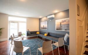 Bright lounge with blue sectional sofa, white chairs, navy rug, wall art, and glass doors.