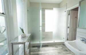 Bright modern bathroom with frosted glass shower, white sink, marble table, and plant.