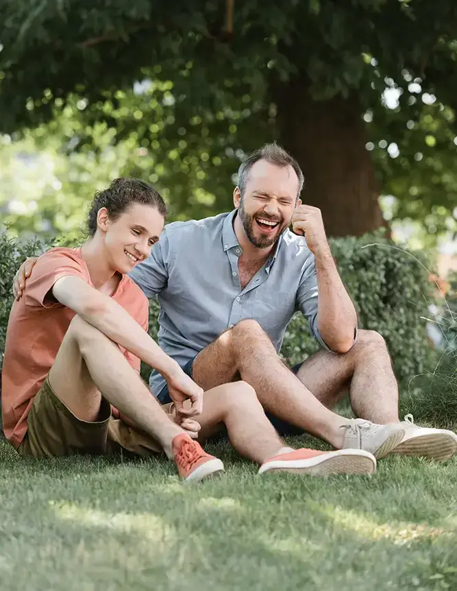 Two men sitting on grass in a park, smiling and laughing together under a tree.