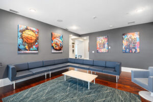 Modern lounge with gray walls, long gray sectional, light wood coffee tables, and colorful sports-themed art.