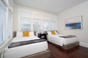 Two modern single beds with white quilts and mustard pillows in a bright room with a shared nightstand and large windows.