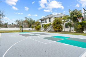 Outdoor basketball court with patterned gray surface, white lines, teal accents, and surrounding building.