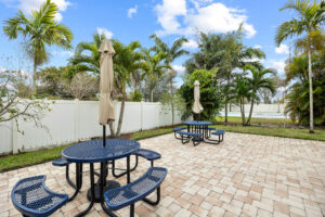 Outdoor patio with blue metal tables and benches, beige umbrellas, and palm trees.