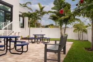 Patio with blue round-top tables, benches, gray bench, brick pavers, and white fence. Trees and lawn.