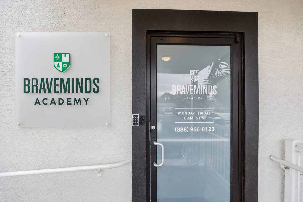 Storefront door with Braveminds Academy sign on beige textured wall, glass door reflection.