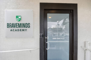 Storefront door with Braveminds Academy sign on beige textured wall, glass door reflection.