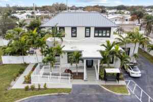 Two-story modern building with gray metal hip roof, white exterior, palm trees, and driveway with car.
