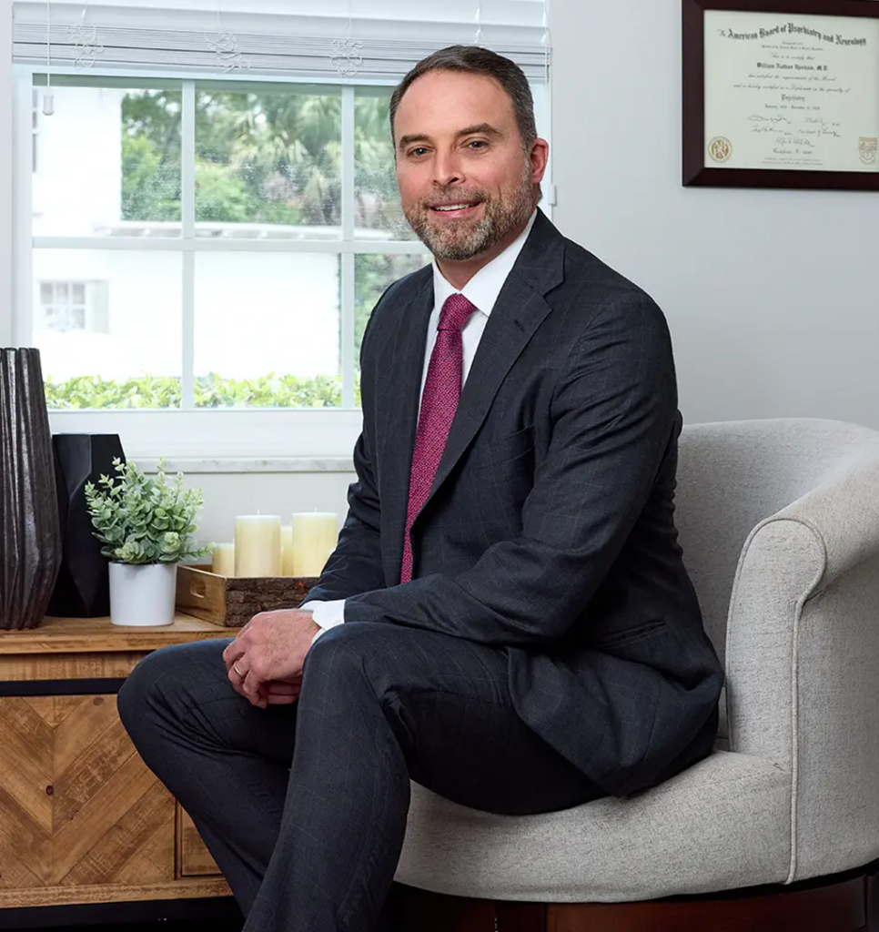 Man in a dark suit and pink tie sits on a light gray armchair in a bright, tidy office.