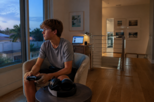 Young man sitting in living room at dusk, holding a game controller, by a large window.